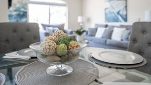 A dining table with a glass bowl containing decorative items, a grey upholstered sofa, and a modern abstract painting in the background. - Luxury Comfort close to Westworld & TPC + Pool&Spa (Scottsdale)