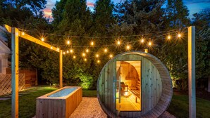A wooden hot tub and a circular wooden structure with a bench, both set on a gravel surface with string lights overhead.