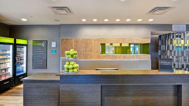 Un'area reception di un hotel dal design moderno, con un banco, un frigorifero fornito di bevande e un cartello che indica 'HOME2' e 'Hilton Honors'.