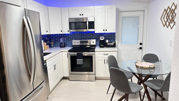 A modern kitchen with white cabinets, a stainless steel refrigerator, a microwave, an oven, and a coffee maker. There is a glass table with chairs and a decorative wall piece.