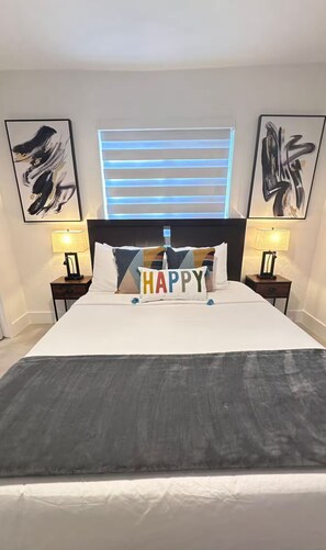 A hotel room with a large bed, two bedside tables with lamps, abstract wall art, a window with horizontal blinds, and a pillow with the word "HAPPY".