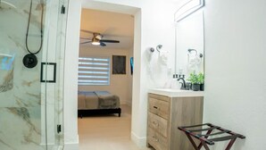 A bathroom with a glass shower enclosure, a wooden vanity with a mirror, and a bed in the adjacent room.