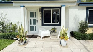 A house with a white exterior, a black-framed window, and a door with the number 812. There are two potted plants on the porch, a white plastic chair, and a small round table with a plant.