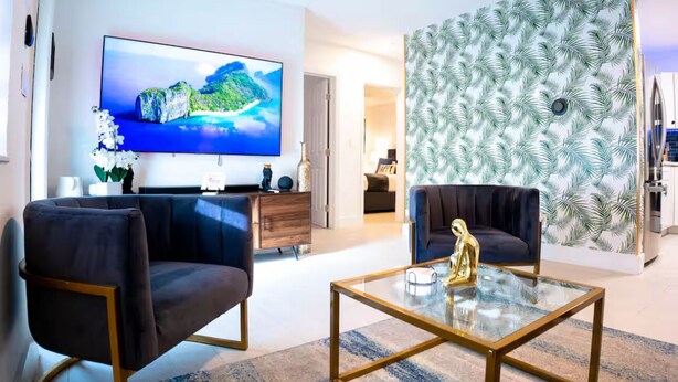 A modern living room with a large flat-screen TV, two dark sofas, a glass coffee table with a golden statue, and a tropical leaf-patterned wall.