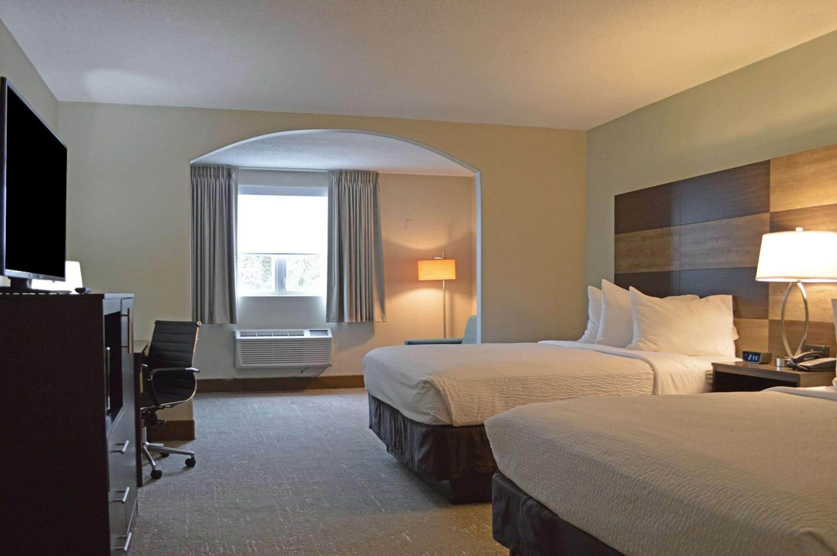 A hotel room with two beds, a TV, a desk, a chair, a lamp, and a window with curtains.