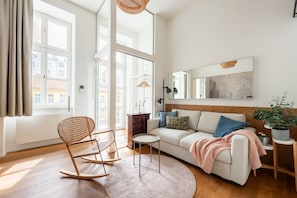 A modern living room with a sofa, a rocking chair, a round table, and a large window with curtains.