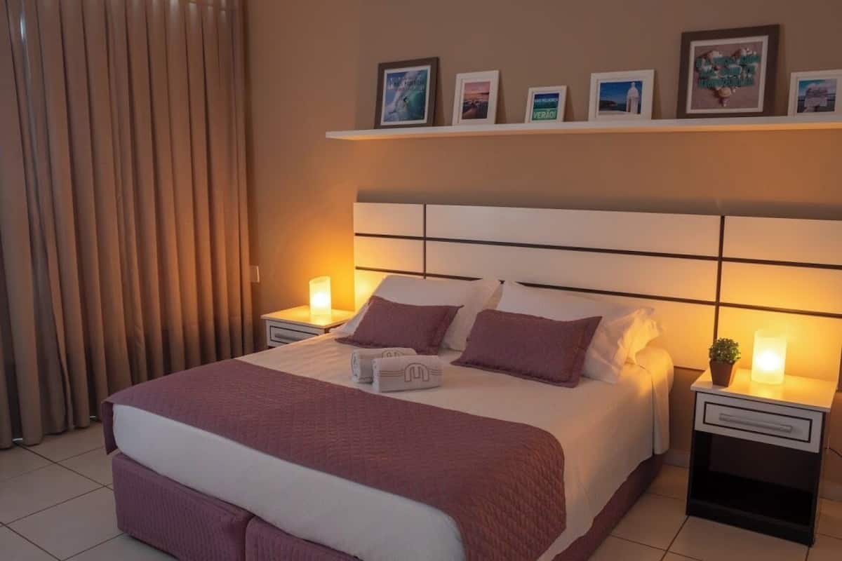 A neatly made bed with a headboard, two bedside tables with lamps, and a shelf above the bed with framed pictures.