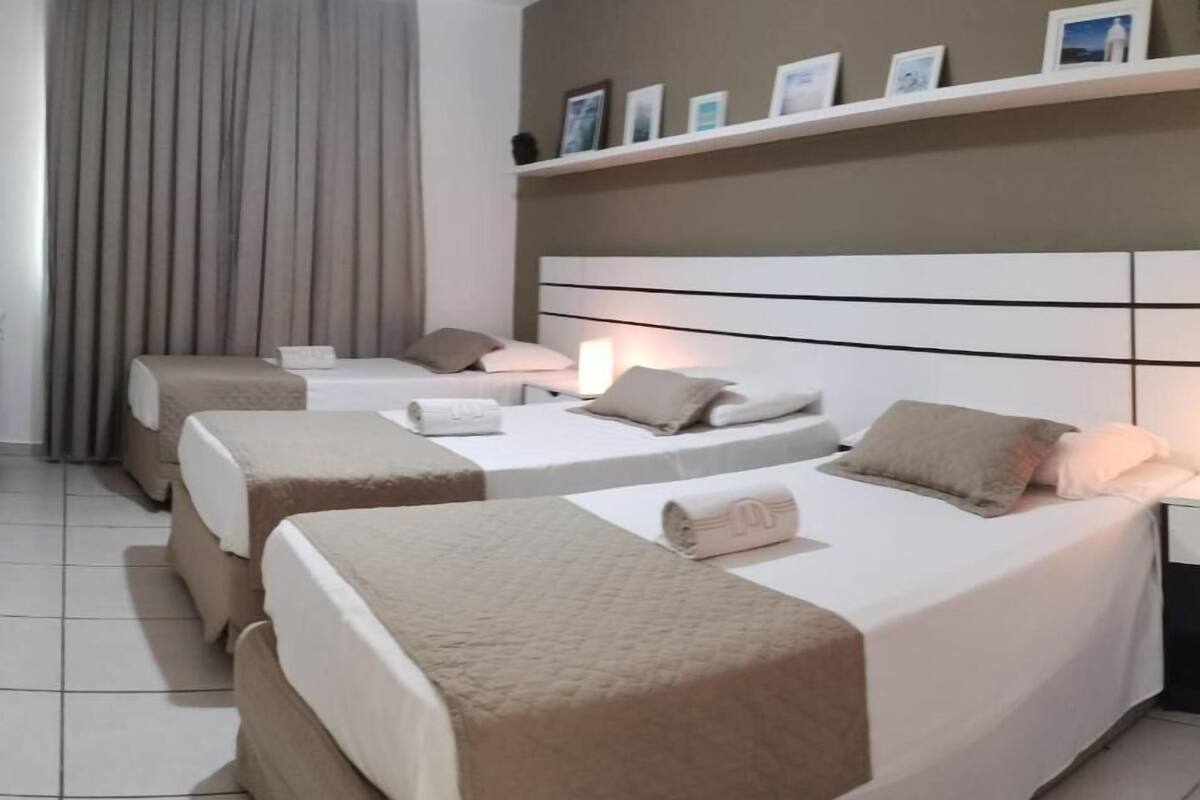 A hotel room with three beds, each with a white and beige bedspread, a headboard, and a bedside lamp.