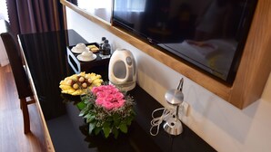 Habitación de hotel con escritorio negro, televisor de pantalla plana en la pared, una mesita con tetera, tazas y bandeja de aperitivos.