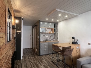 A compact kitchen with wooden walls, a herringbone-patterned backsplash, and modern appliances. There is a wooden dining table with two chairs and a small black lamp on the countertop.
