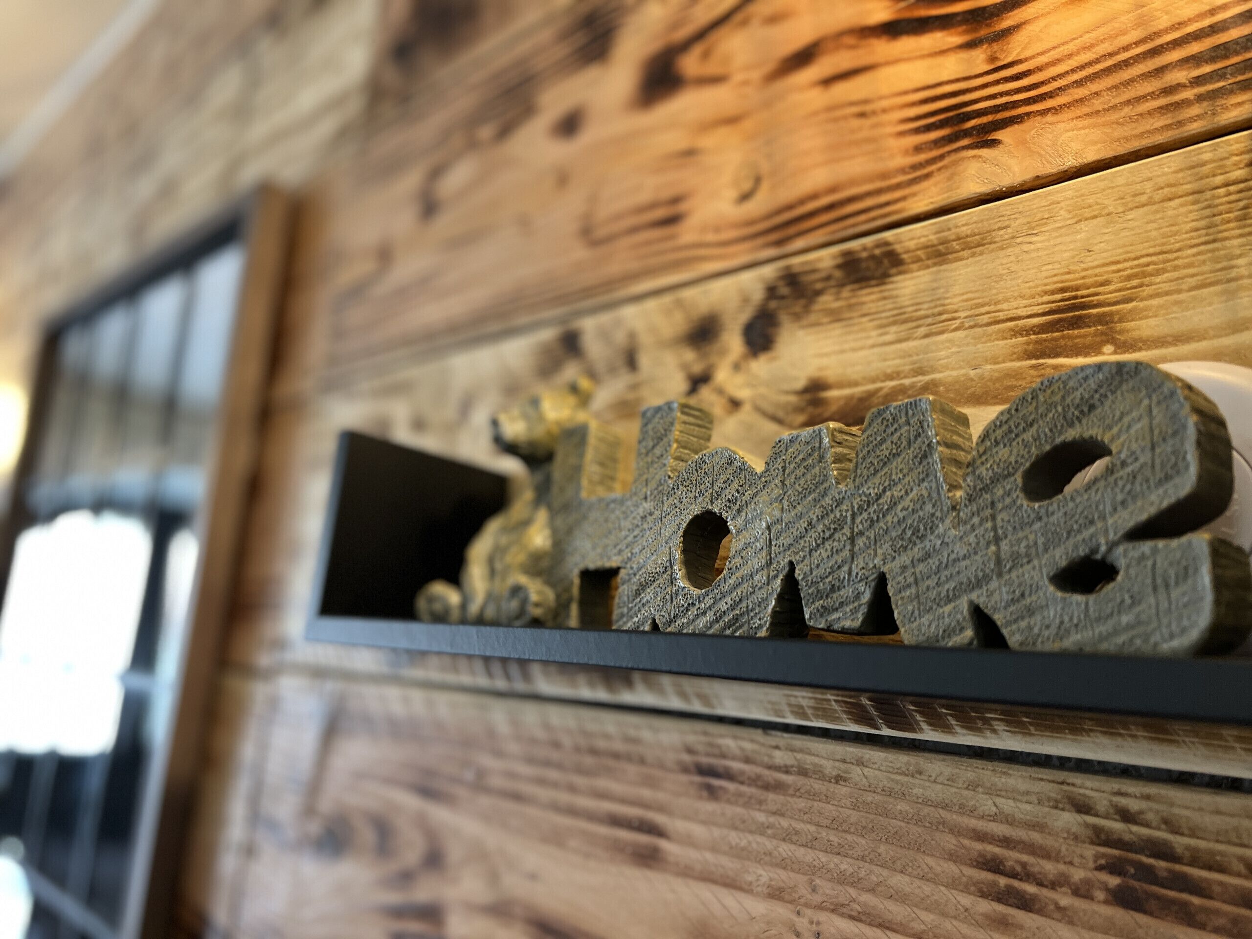 Puinen seinä, jossa on hylly, ja hyllyllä on koristeellisella kirjasintyypillä kirjoitettu sana ”Home” (koti).
