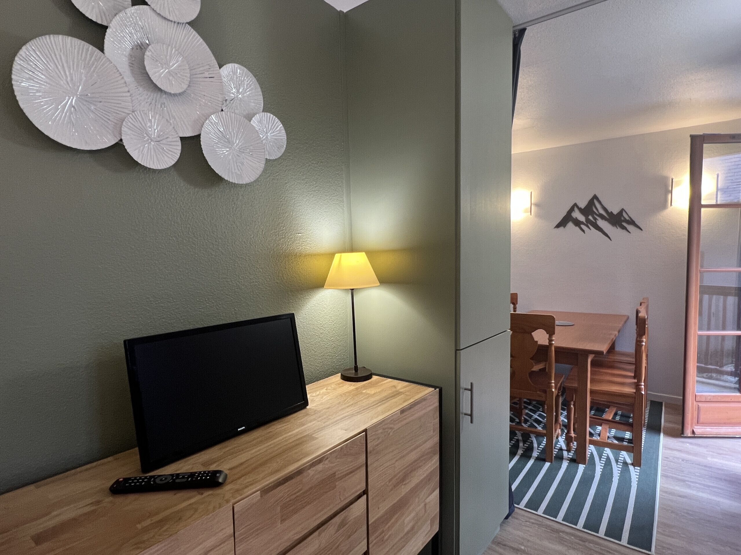 A room with a TV on a wooden cabinet, a lamp, a dining table with chairs, and a wall with a mountain decoration.