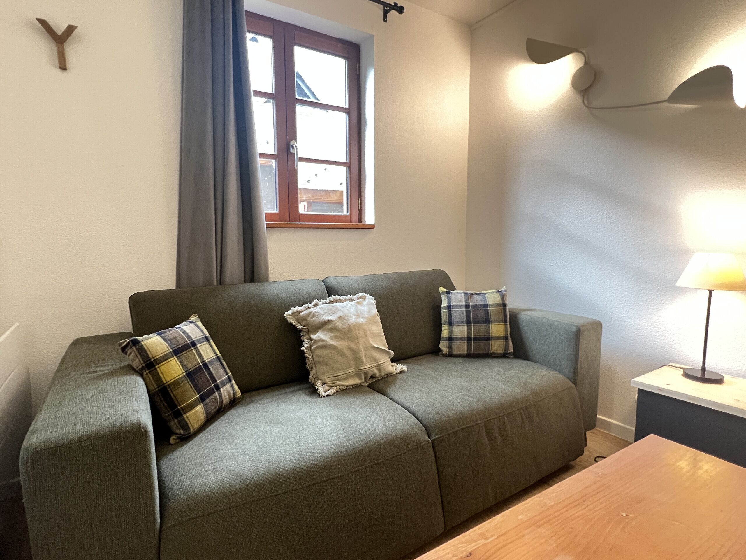 A living room with a sofa, two pillows, a wooden table, a lamp, a window with curtains, and a wall-mounted light fixture.