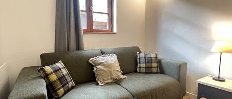 A living room with a sofa, two pillows, a wooden table, a lamp, a window with curtains, and a wall-mounted light fixture.