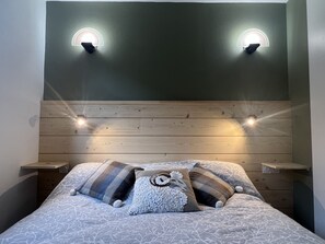 Uma cama bem arrumada com roupa de cama estampada e almofadas decorativas, ladeada por mesas de cabeceira de madeira e duas luminárias fixadas na parede.