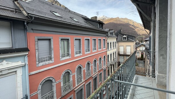 Una calle estrecha en la ciudad con edificios que tienen ventanas en arco y balcones, un techo empinado y un telón de fondo montañoso.