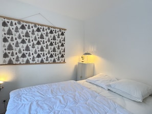 A bedroom with a bed, white bedding, a nightstand with a lamp, and a wall-mounted picture of trees and leaves. - -Centre ville, T2 avec terrasse et parking couvert. (Cauterets)