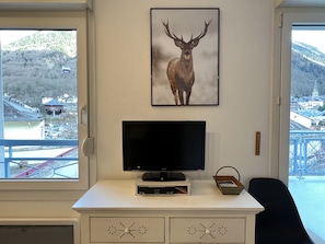 A room with a TV on a white cabinet, a framed deer picture on the wall, and a view of a town and mountains through the window. - -Centre ville, T2 avec terrasse et parking couvert. (Cauterets)
