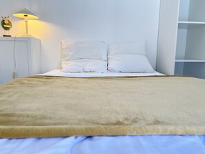 Cama bem arrumada com manta bege, travesseiros brancos e abajur com um objeto decorativo na mesa de cabeceira.