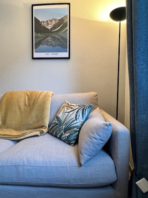 Ein Zimmer mit einem hellblauen Sofa, einem Kissen mit tropischem Blattmuster, einer beigen Tagesdecke und einem gerahmten Kunstwerk an der Wand.