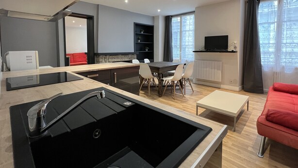 A modern kitchen with a black stove, a dining area with a wooden table and chairs, a red sofa, and a television on a stand.