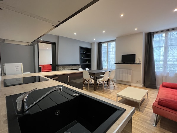 A modern kitchen with a black sink, dining area with white chairs, a red sofa, and a TV on a shelf.