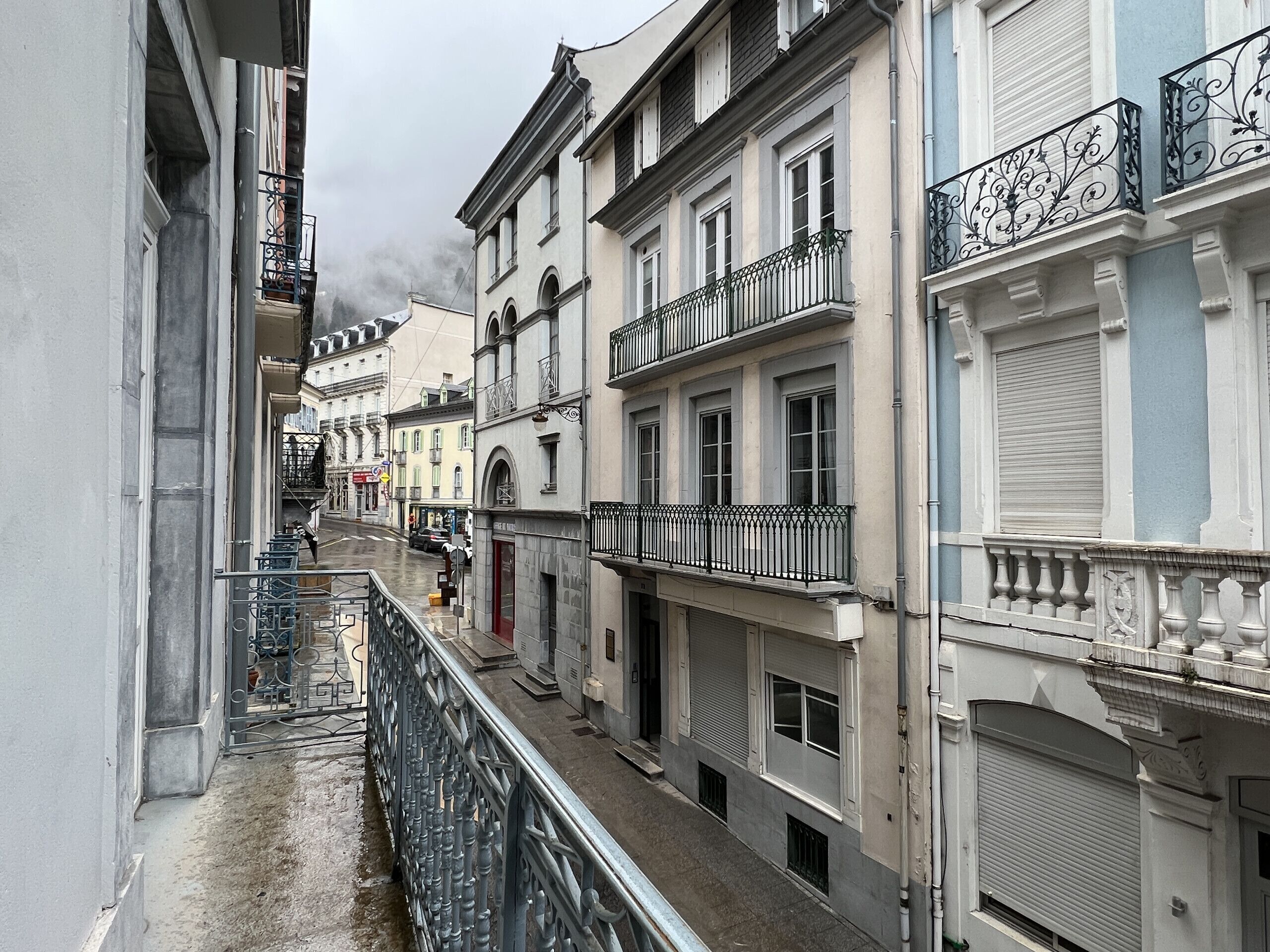 A narrow street with buildings on either side, featuring balconies with metal railings and a variety of architectural styles.