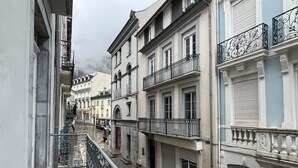 A narrow street with buildings on both sides, featuring balconies with wrought iron railings and shutters on the windows.