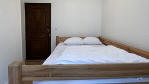 Una cama individual con estructura de madera y ropa de cama blanca en una habitación con paredes blancas y una puerta de madera.
