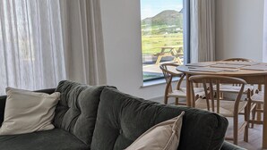 Een moderne woonkamer met een donkergroene bank, beige kussens en een eethoek met een houten tafel en stoelen. Een groot raam biedt uitzicht op een groen landschap.