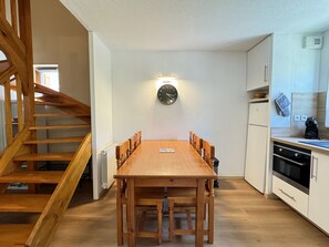A wooden dining table with chairs, a clock on the wall, and a kitchen area with cabinets and appliances. - Duplex 40m2. Balcon. Parking. 5P (Cauterets)