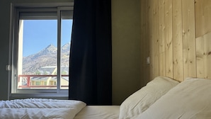 A bedroom with a wooden headboard, a large window with a view of mountains, and a bed with white bedding. - Joli 3 pièces 40m2. 2 chambres (Cauterets)
