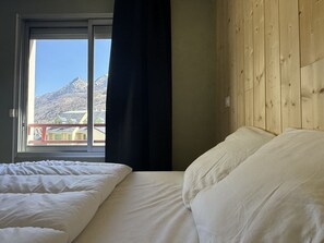 A bedroom with a wooden headboard, a large window with a view of mountains, and a bed with white bedding.