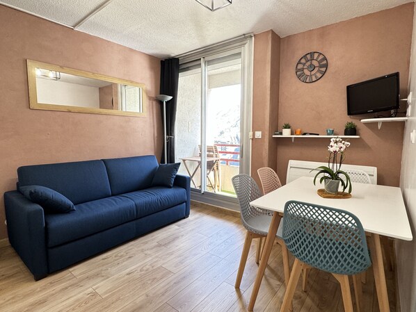 A furnished living room with a sofa, chairs, a dining table, a TV, and a window with a view.