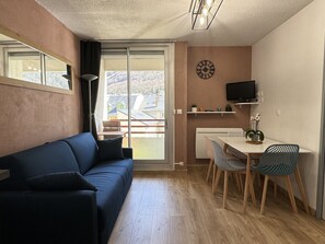 A compact living space with a sofa, a dining table with chairs, a TV, and a view of buildings through a window. - Joli 3 pièces 40m2. 2 chambres (Cauterets)