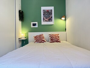 A neatly made bed with white bedding, two decorative pillows, a framed poster on the wall, a bedside lamp, and a small nightstand.