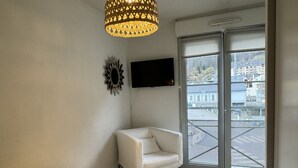 Une chambre à coucher comprenant un lit, une chaise, un téléviseur fixé au mur et une fenêtre donnant sur des immeubles.