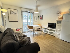 A modern living room with a sofa, a dining table, and chairs. There is a television and a refrigerator in the kitchen area.