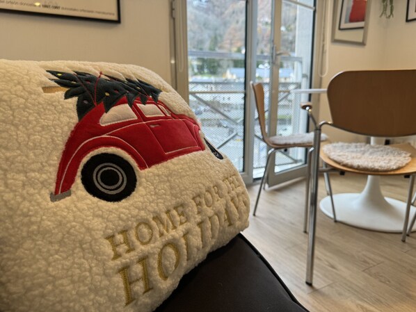 A plush pillow with a car and the text "HOME FOR HONEY" on a couch in a modern room with a chair and a view of a balcony.