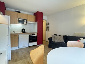 A compact kitchen with a microwave, oven, and dishwasher. A black sofa and a wooden chair are in the living area. - 100m télécabine ou commerces. 4P (Cauterets)