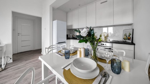 A modern kitchen with white cabinets, a dining table set with plates, glasses, and a vase of flowers. - Śródmieście Apartment - 8th Floor, Workspace, Tram - by Rentujemy (Warszawa)