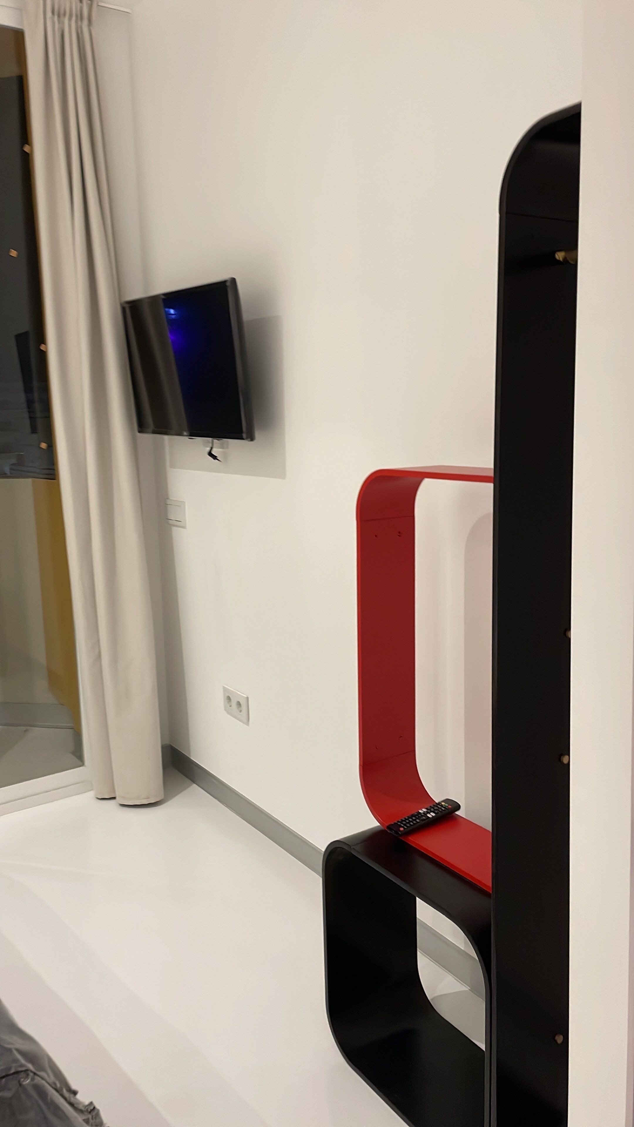 A modern room with a flat-screen TV mounted on the wall, a red and black chair, and a window with curtains.