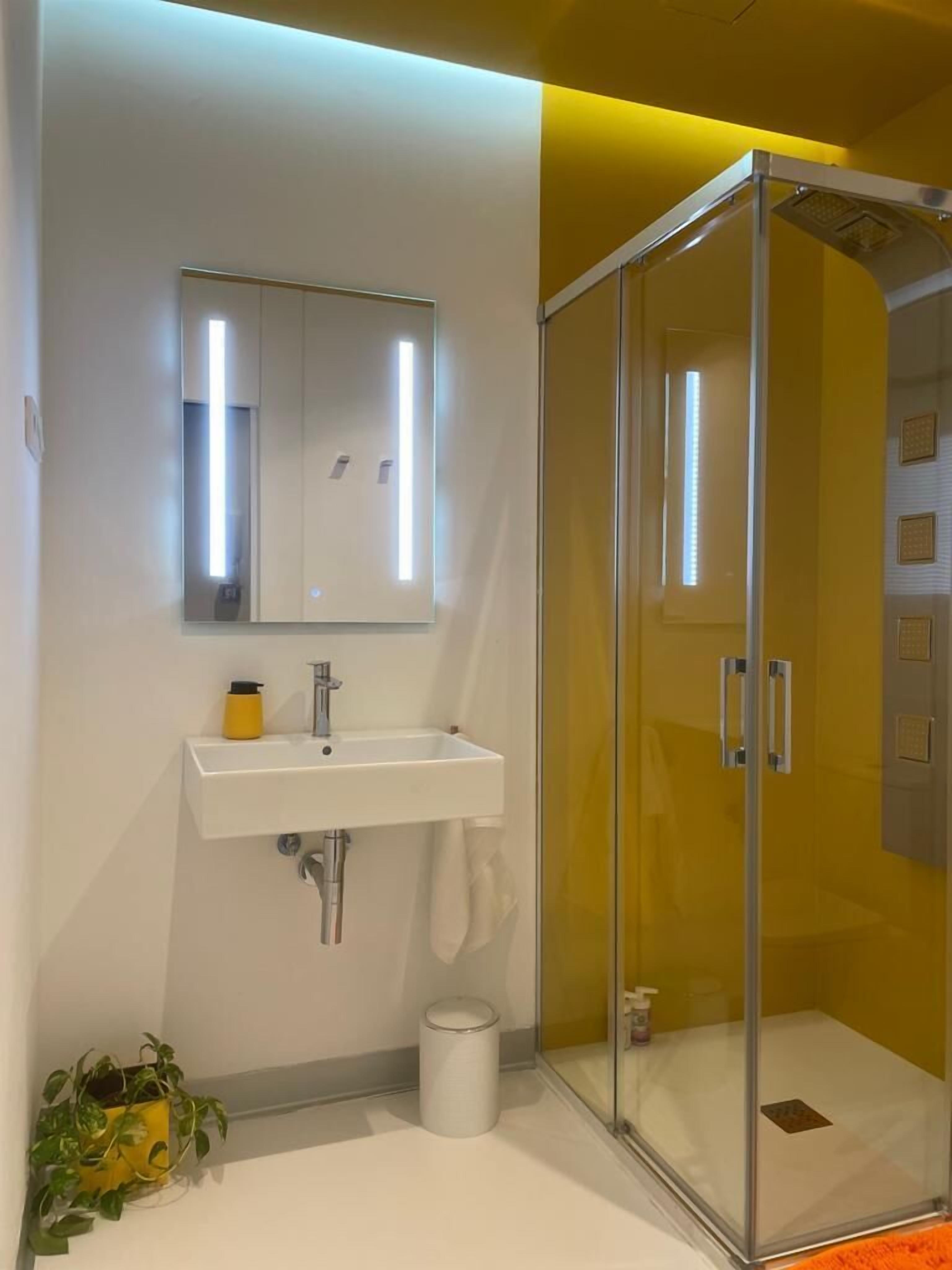 Un baño moderno con ducha de vidrio, un lavamanos blanco con espejo y una pequeña planta en maceta en el suelo.