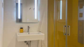 Un baño moderno con ducha de vidrio, un lavamanos blanco con espejo y una pequeña planta en maceta en el suelo.