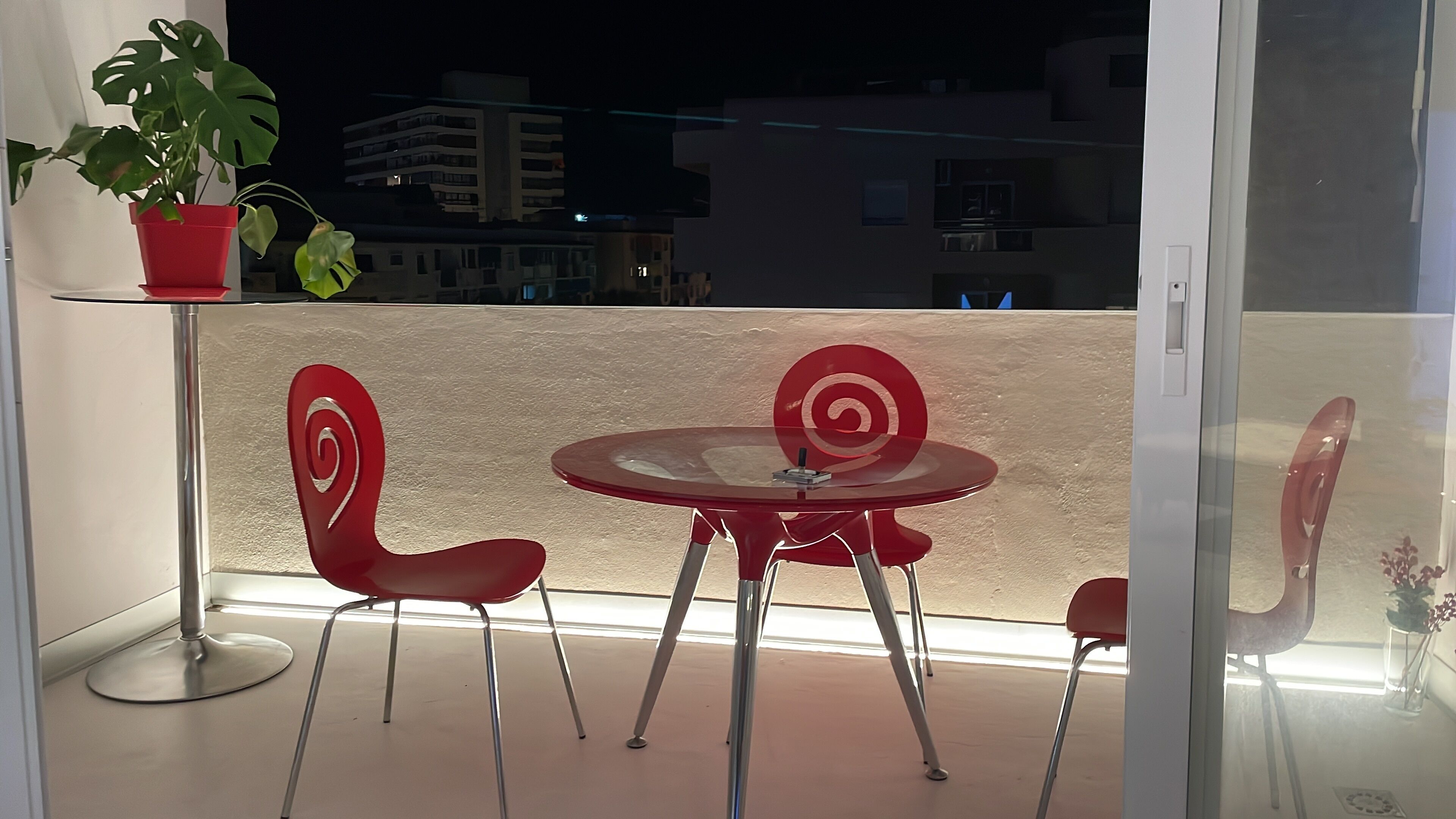 Un balcon avec une table en verre et deux chaises rouges, un pot de fleurs et une vue sur les bâtiments la nuit.