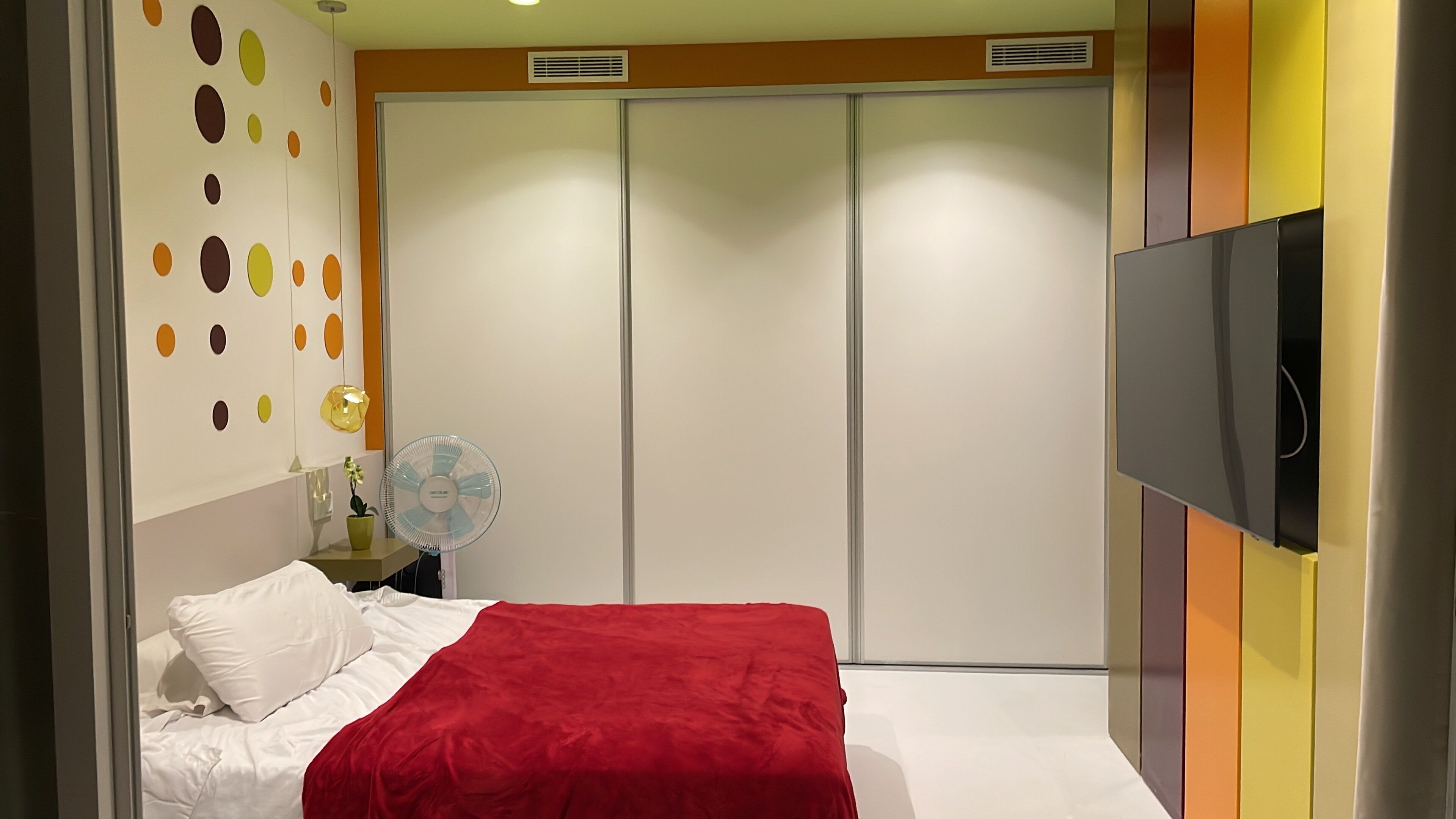 A bedroom with a bed, a red blanket, a wall-mounted TV, a fan, and a wardrobe.