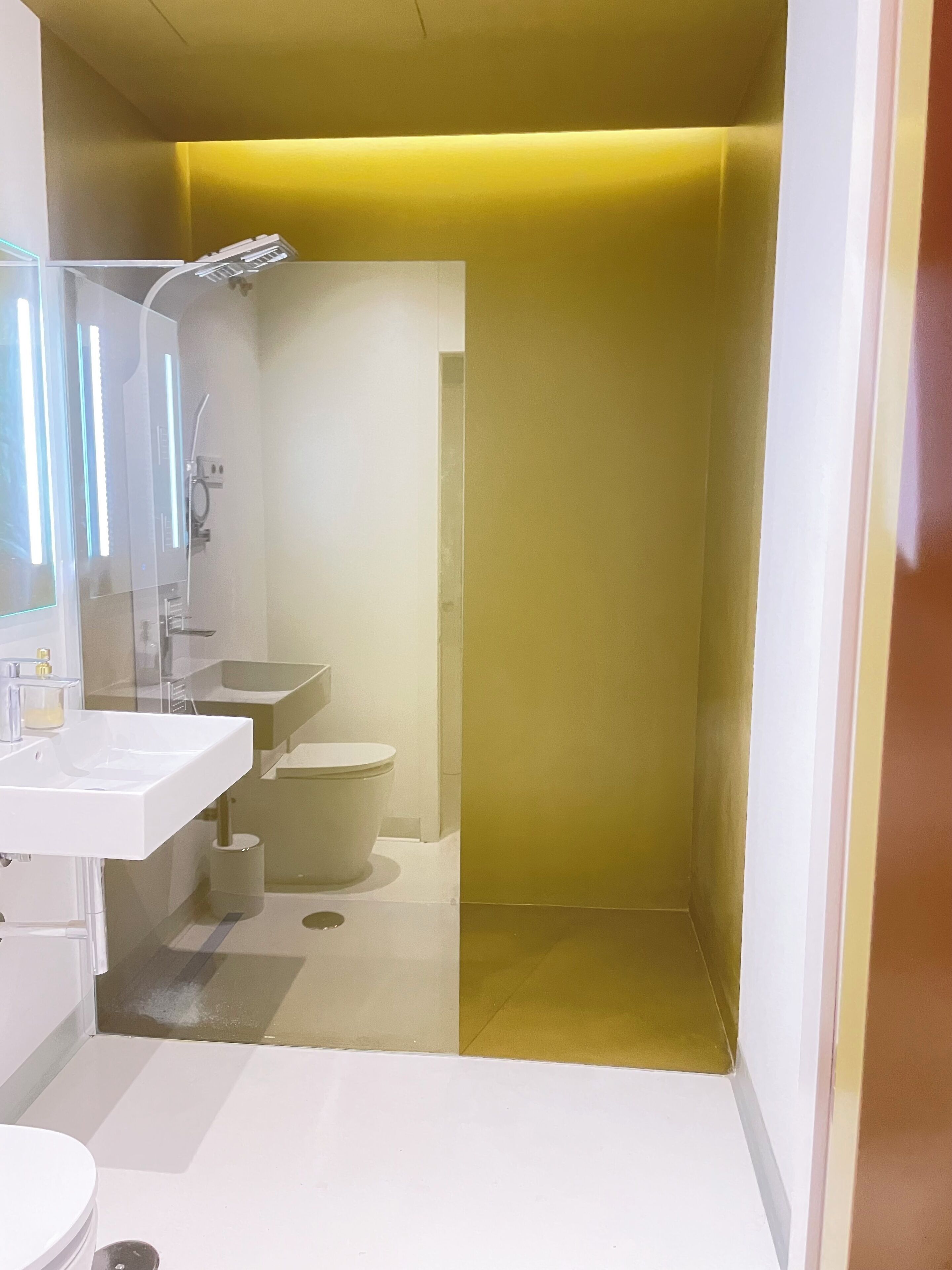 A modern bathroom with a white sink, mirror, and toilet. There is a shower area with a glass door. The walls are painted in a light color, and the floor is tiled.
