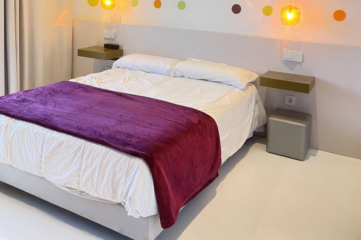 A modern hotel room with a large bed, white bedding, a purple throw blanket, and a wall decorated with colorful circular patterns.