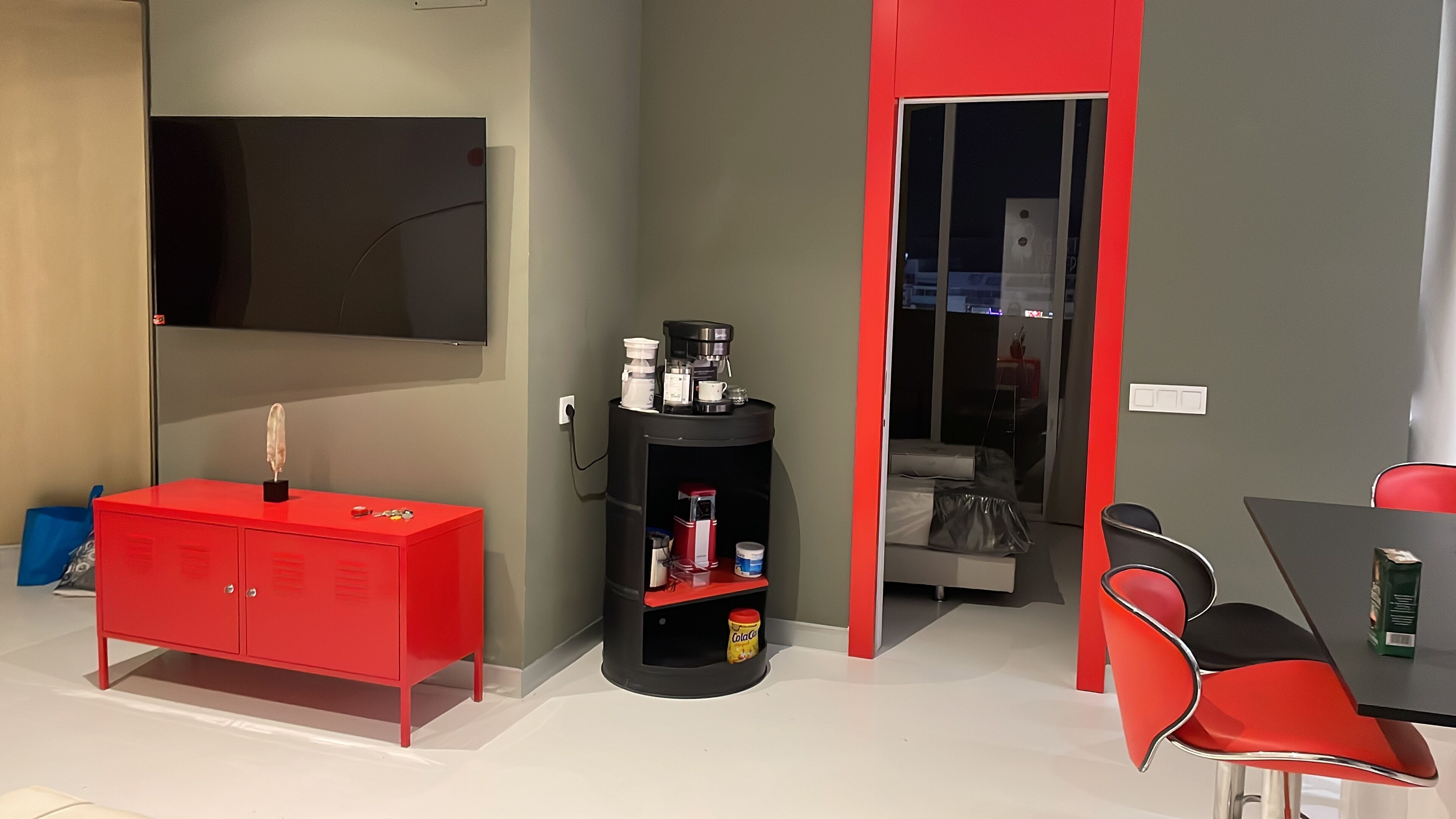 A compact living space with a red cabinet, a black coffee station, a television, and a dining area with red chairs.