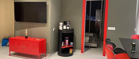 A compact living space with a red cabinet, a black coffee station, a television, and a dining area with red chairs.